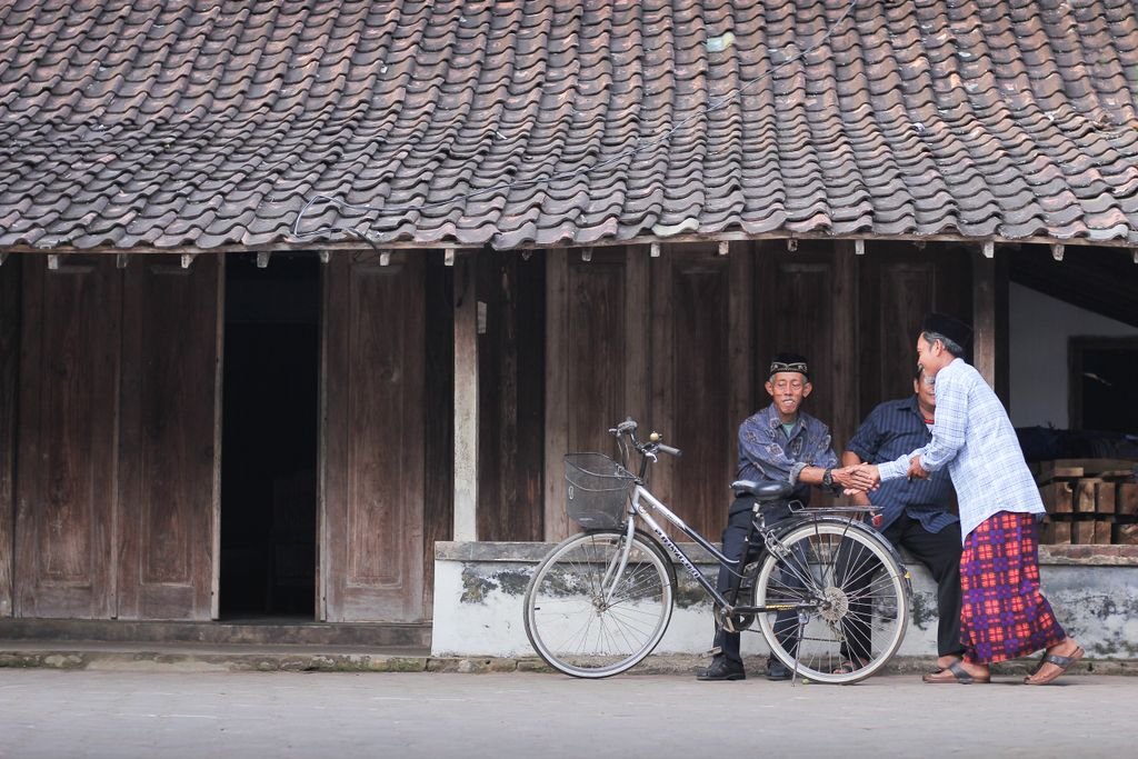 Elderly men shake hands outside a rustic house with a vintage bicycle in view.